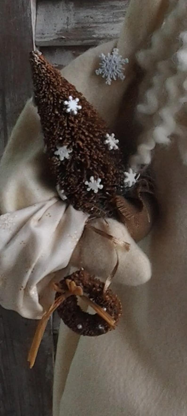 May include: A white fabric Santa Claus figure with a brown, twig-like Christmas tree in its hand. The tree is decorated with white snowflake ornaments.
