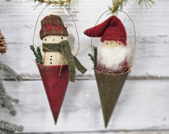 May include: Two felt Christmas decorations hanging from a white wooden background. The decoration on the left is a snowman with a red and green plaid hat and a green scarf. The decoration on the right is a gnome with a red hat and a white beard. Both decorations are in cone-shaped containers.