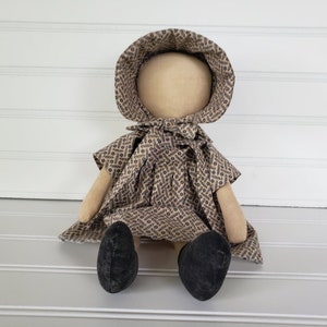 May include: A handmade rag doll with a brown and blue patterned dress and a matching bonnet. The doll is sitting on a white surface.