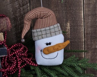 May include: A handmade snowman ornament with a carrot nose, black eyes, and a stitched smile. The snowman wears a brown and tan striped hat with a plaid brim. The ornament is displayed on a bed of evergreen branches.