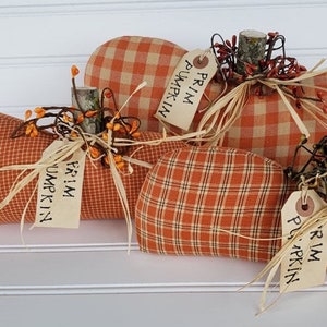May include: Three orange and white plaid fabric pumpkins with brown twig stems and green berry sprigs. Each pumpkin has a tag that reads "Prim Pumpkin".