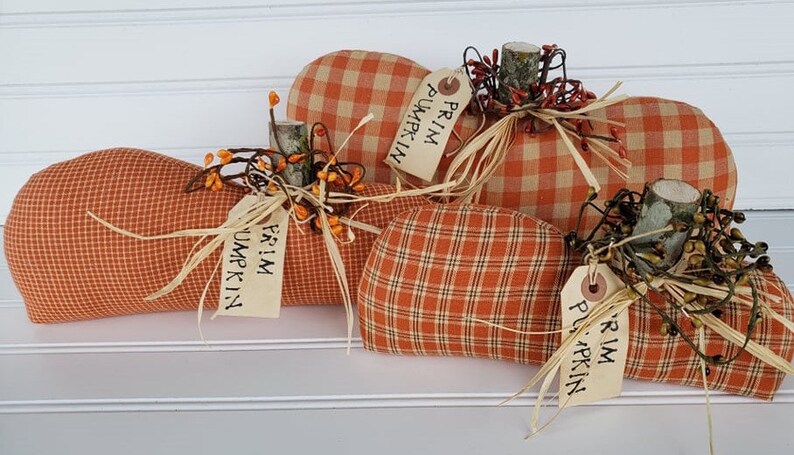 May include: Three orange and white plaid fabric pumpkins with brown twig stems and green berry sprigs. Each pumpkin has a tag that reads "Prim Pumpkin".