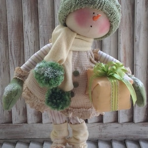 Può includere: Una bambola di pupazzo di neve fatta a mano con un cappello di lana verde, una sciarpa crema e un vestito a righe. La bambola tiene in mano un regalo avvolto con un nastro verde. La bambola è in piedi su una superficie di legno.