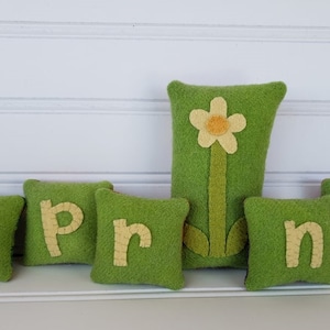Op de afbeelding: Groene viltkussens met het woord "Spring" gespeld in gele viltletters. Een groen viltkussen met een gele viltbloem bevindt zich in het midden.