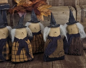 May include: Five handmade witch dolls with black pointed hats, white hair, and plaid skirts. Each doll has a dark blue scarf and a tan body. The dolls are arranged on a wooden surface, evoking a rustic, autumnal theme.