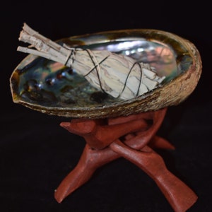 May include: A white sage smudge stick bundled with twine sits in a abalone shell on a wooden stand. The shell has iridescent colors of blue, green, and purple.
