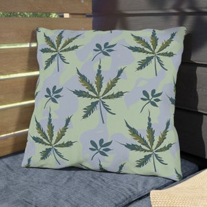 Puede incluir: Una almohada de color verde claro con un patrón repetitivo de hojas estilizadas de color verde oscuro y marrón. La almohada está sobre una superficie texturizada azul y gris.