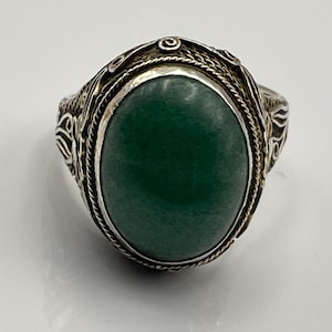 May include: A silver ring featuring a large, oval-shaped, dark green stone set in the center. The metal band is decorated with intricate, raised filigree patterns and twisted wire detailing surrounding the bezel of the smooth, polished gemstone.
