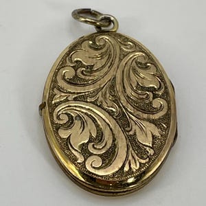 May include: An oval-shaped gold-toned metal locket pendant featuring an intricate embossed scrollwork pattern across the front surface. The design includes raised leaf-like motifs set against a textured background, complete with a small circular bail at the top.