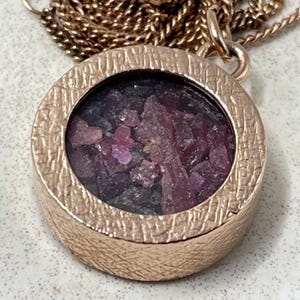 May include: A round rose gold pendant necklace featuring a textured metal frame and a circular centre filled with small, irregular, deep pink and dark purple gemstone fragments. The pendant hangs from a matching rose gold chain link necklace.