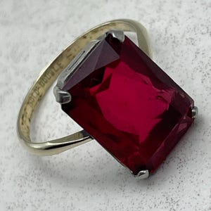 May include: A gold-tone ring featuring a large, rectangular, faceted deep red stone held by four silver-tone prongs. The inner band is clearly engraved with the text "DOUBLÉ" and "GOLD". The design showcases a classic setting with a polished finish.