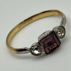 Könnte beinhalten: Ein goldfarbener Ring mit der Innengravur "BAITED GOLD". Die Fassung besteht aus einem quadratischen violetten Stein in der Mitte, der von zwei kleineren, runden, klaren Steinen flankiert wird, alle in einer silberfarbenen Metallfassung.