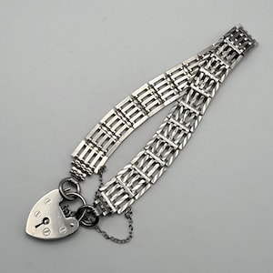 Könnte beinhalten: Ein silberfarbenes Metallarmband mit einem mehrgliedrigen Ketten-Design und strukturierter Oberfläche. Das Armband wird mit einem herzförmigen Vorhängeschloss-Anhänger gesichert, der ein zentrales Schlüsselloch und dekorative Schraubendetails aufweist.