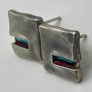 Puede incluir: Un par de pendientes de botón de plata con una forma cuadrada irregular y texturizada. Cada pendiente presenta un recorte rectangular que contiene una incrustación horizontal de esmalte turquesa vibrante y rojo intenso, con un poste.