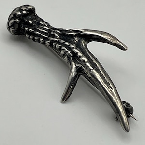 May include: A silver-toned brooch shaped like a textured deer antler. The metal piece features a dark, oxidised finish in the crevices to highlight the organic, rugged surface details and the pointed, branching structure of the antler design.