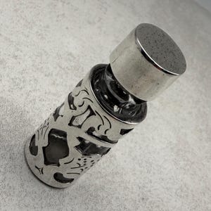Può includere: Un flacone di profumo color argento con un design traforato ornato. Il flacone ha un corpo cilindrico e un tappo. L'intricato motivo avvolge il flacone, creando un effetto decorativo. Il tappo ha una forma arrotondata.