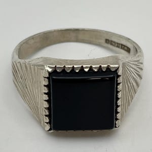 May include: A silver ring featuring a square, polished black stone set in a scalloped bezel. The band displays a textured, sunburst-like engraved pattern radiating from the central setting, with visible hallmarks stamped on the inner surface.