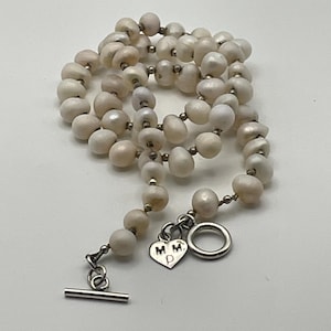 May include: A necklace featuring irregular white pearls interspersed with small silver-toned beads. The piece includes a silver-toned toggle clasp and a heart-shaped charm engraved with the letters M, M, and P on the front surface.