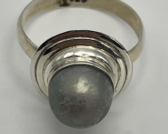 Puede incluir: Un anillo de plata con una piedra gris, lisa y redonda, montada sobre una base de metal pulido. El interior de la banda tiene grabado el sello 925, que indica una composición de plata de ley. El diseño incluye capas metálicas circulares.