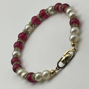 May include: A beaded bracelet featuring alternating round white pearls and translucent deep pink beads. Each bead is separated by a small, thin gold-toned spacer ring encrusted with tiny clear crystals. The bracelet includes a gold-toned metal clasp.