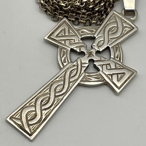 May include: A silver-toned metal Celtic cross pendant featuring intricate embossed knotwork patterns throughout the arms and center circle. The cross is suspended from a thick, round silver-toned metal chain link necklace.
