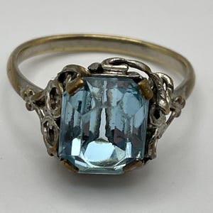 Pode incluir: Um anel com uma pedra preciosa retangular azul-clara de corte esmeralda montada numa banda metálica. O engaste é decorado com trabalhos metálicos intrincados e orgânicos em forma de videira que rodeiam a pedra central da peça.