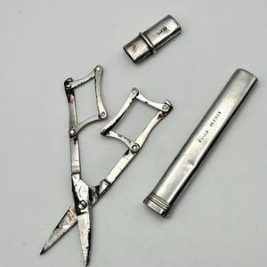 May include: A set of silver-toned folding scissors with a collapsible lattice handle mechanism, accompanied by a matching cylindrical metal case and a separate small cap. The metal surfaces show signs of wear and include visible hallmarks on the case.