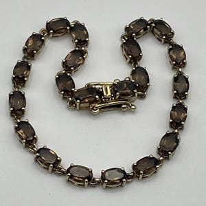 Peut inclure: Un bracelet composé d'une série de pierres précieuses quartz fumé marron translucides de forme ovale, serties dans une monture métallique. Chaque pierre est reliée à la suivante par un maillon, avec un fermoir boîte rectangulaire en métal.