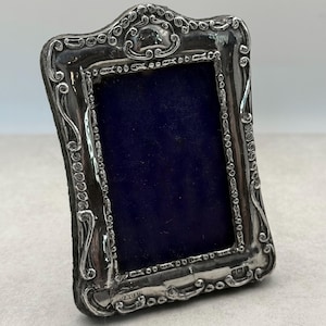 May include: A rectangular silver picture frame featuring an ornate, embossed border with scrolling patterns and a deep blue velvet backing. The frame has a polished metallic finish and includes small engraved hallmarks at the bottom edge.