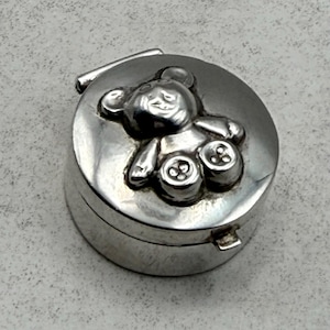 Pode incluir: Uma pequena caixa redonda de metal prateado com tampa articulada. O topo da tampa apresenta um design em relevo de um urso de peluche com detalhes em forma de botões nos pés, sobre uma superfície metálica lisa e refletora.