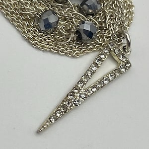 May include: A silver-toned necklace featuring a delicate chain adorned with dark gray faceted beads and a triangular pendant encrusted with small clear crystals. The pendant has a sharp, pointed design and is attached to the chain with a metal jump ring.