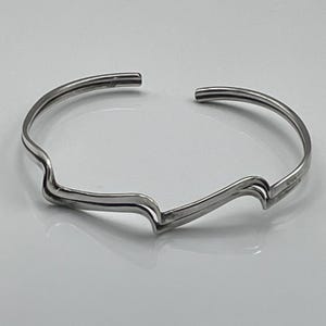 May include: A silver-toned metal cuff bracelet featuring a continuous open-ended band with a distinct double-layered wave or mountain-like silhouette. The polished surface reflects light, highlighting the smooth, curved lines of the jewellery piece.