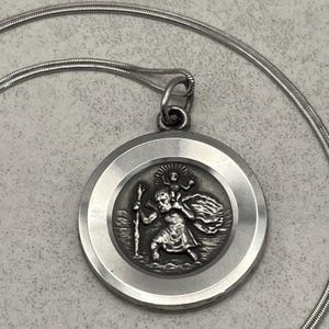 May include: A silver-toned pendant necklace featuring a round medallion with a detailed relief of Saint Christopher carrying a child. The pendant is suspended from a thin, silver chain. The medallion has a raised border.