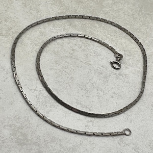 Könnte beinhalten: Eine silberfarbene Metallkettenhalskette mit einer Reihe kleiner, rechteckiger, ineinandergreifender Glieder. Die Halskette ist in einer kreisförmigen Spirale angeordnet und verfügt an einem Ende über einen runden Federringverschluss.
