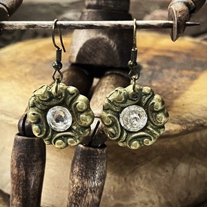 May include: A pair of gold-toned earrings with a detailed floral design and a clear crystal in the center of each earring.