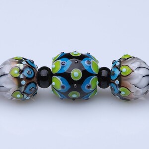May include: A strand of colorful glass beads with black accents. The beads are round and have a variety of patterns, including polka dots, swirls, and geometric shapes. The beads are strung on a black cord and are separated by small, clear beads.