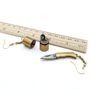 Könnte beinhalten: Ein Paar goldene Ohrringe mit einem kleinen Taschenmesser und einem Feuerzeug-Anhänger. Die Ohrringe hängen an einem Lineal aus Holz, das in Zoll eingeteilt ist.