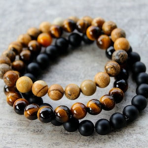 May include: Three beaded bracelets stacked on top of each other. The top bracelet is made of light brown beads with dark brown streaks. The middle bracelet is made of tiger's eye beads. The bottom bracelet is made of black beads.