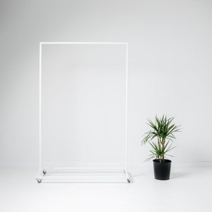Peut inclure: Un portant à vêtements en métal blanc sur roulettes est placé dans une pièce lumineuse. Une plante verte en pot avec de longues feuilles fines se trouve à droite. Le portant a un cadre rectangulaire et une étagère inférieure.
