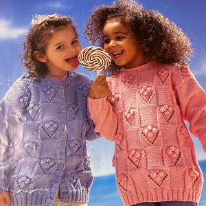 Op de afbeelding: Twee jonge meisjes dragen gebreide vesten met hartvormige ontwerpen. Het meisje links draagt een lavendelkleurig vest en het meisje rechts draagt een roze vest. Ze lachen allebei en houden een lolly.