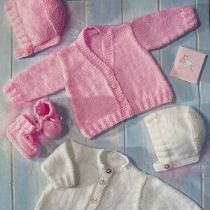May include: Two knitted baby sets, one pink and one white. Each set includes a cardigan, hat and booties. The pink set has a button fastening and the white set has a button fastening with decorative embellishments.