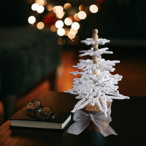 Könnte beinhalten: Ein weißer, gehäkelter Schneeflocken-Weihnachtsbaumschmuck mit einer Jutesockel und einer weißen Schleife. Der Schmuck liegt auf einem Holztisch mit einem Buch und einer Brille.