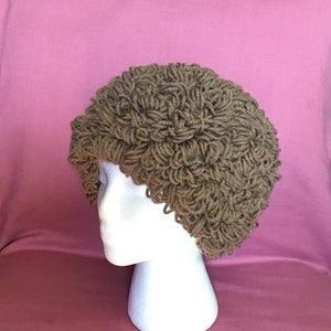 Może przedstawiać: Brązowy, dziergany na drutach kapelusz o kręconej fakturze. Kapelusz znajduje się na białej głowie manekina.