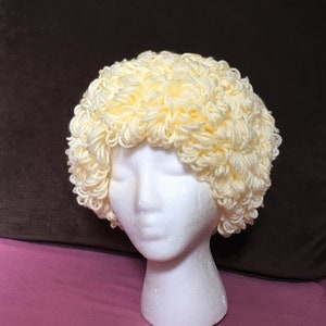 Puede incluir: Una peluca de ganchillo amarilla en una cabeza de maniquí blanca. La peluca está peinada en forma de afro rizado y voluminoso.