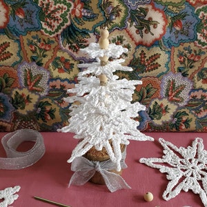 May include: White crocheted snowflake ornaments, a white yarn ball, wooden beads, and a crochet hook on a pink surface. A white crocheted Christmas tree with wooden beads is in the background.