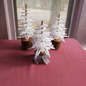 May include: Three decorative white crochet Christmas trees with wooden bases and silver bows. The trees are arranged on a red tablecloth in front of a window with a view of bare trees.