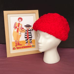 May include: A bright red, chunky knit hat is displayed on a white mannequin head. Beside it is a framed vintage advertisement featuring Ronald McDonald and the Hamburglar. The advertisement has the text "ROWALD & HAMBURGIAR".