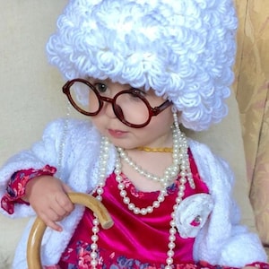 Peut inclure: Un jeune enfant déguisé en femme âgée. Il porte un chapeau en crochet blanc, une robe rose à imprimé floral, un cardigan blanc, un collier de perles et des lunettes rondes. Il tient une canne en bois.