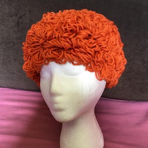 May include: An orange, handmade, crocheted hat with a curly texture. The hat is displayed on a white foam mannequin head. The hat's color is a vibrant, warm orange, and the yarn appears thick and textured.