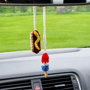 May include: Crocheted car accessories hanging from a rearview mirror. One is a hot dog with mustard, the other is a red, white, and blue popsicle. Both are suspended by white cords.